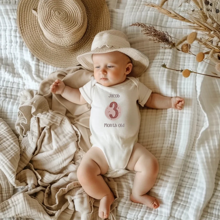 Baby wearing pastel romper with '3 Month Old' print, with a hat, on a cozy blanket backdrop.