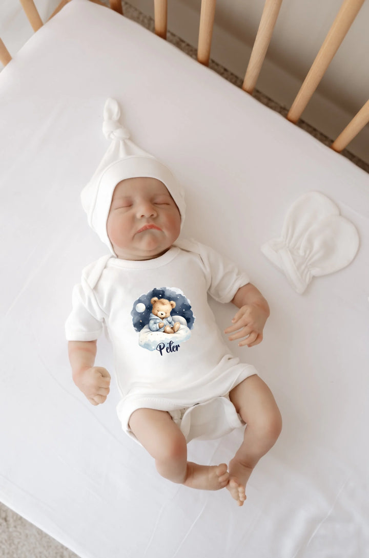 Baby wearing personalized romper with teddy bear design and blue clouds, lying on a crib mattress.