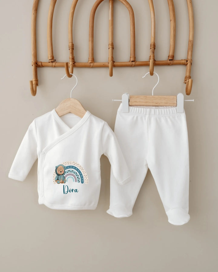 Gepersonaliseerde babykledingset met naam en regenboogprint, bestaande uit een wit jasje en broekje.