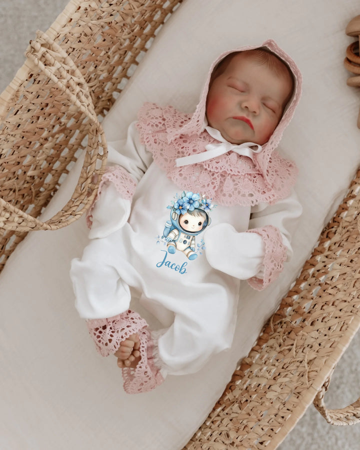 Newborn baby girl wearing a personalized white onesie with pink lace hood and cute astronaut print, lying in a cozy basket.
