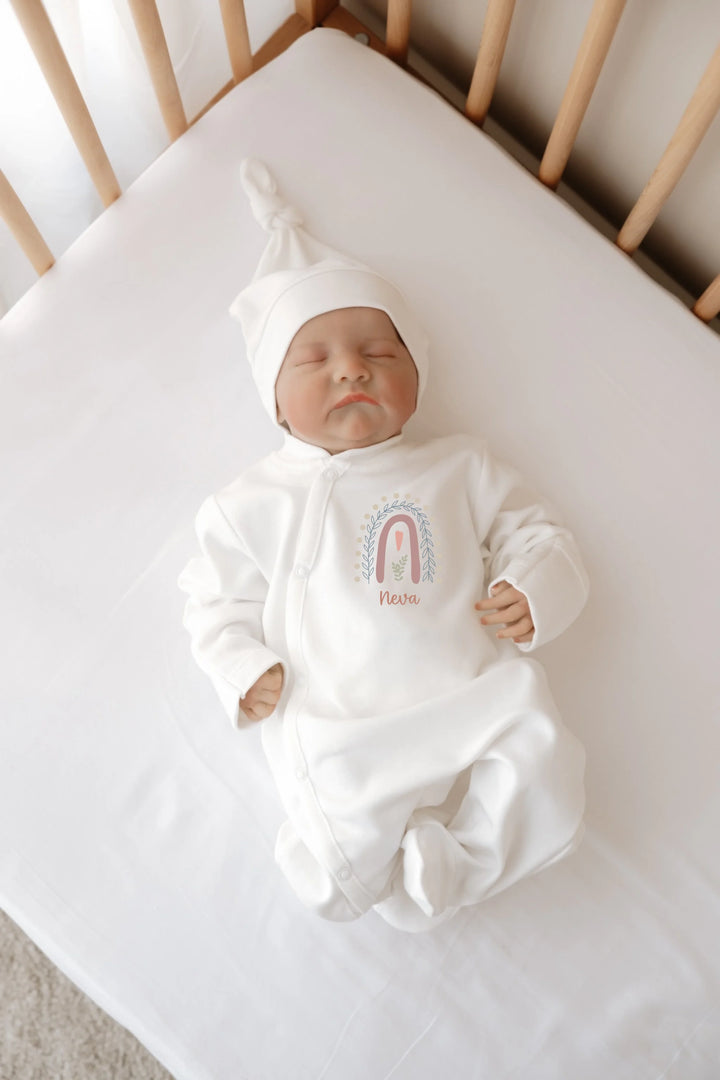 Newborn baby in soft white onesie with rainbow design and personalized name, lying in crib.