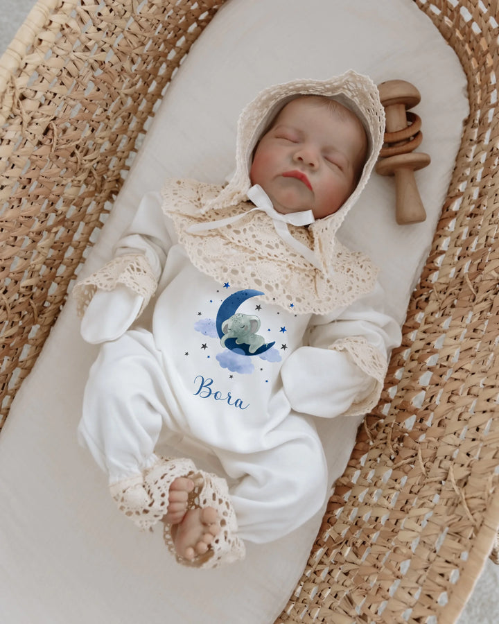 Newborn baby wearing a personalized onesie with lace, featuring a sleeping elephant under the moon, in a cozy basket.