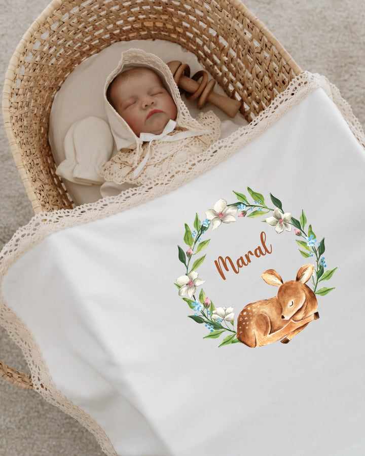 Baby sleeping in a basket, covered with a personalized blanket featuring a deer and floral wreath design.