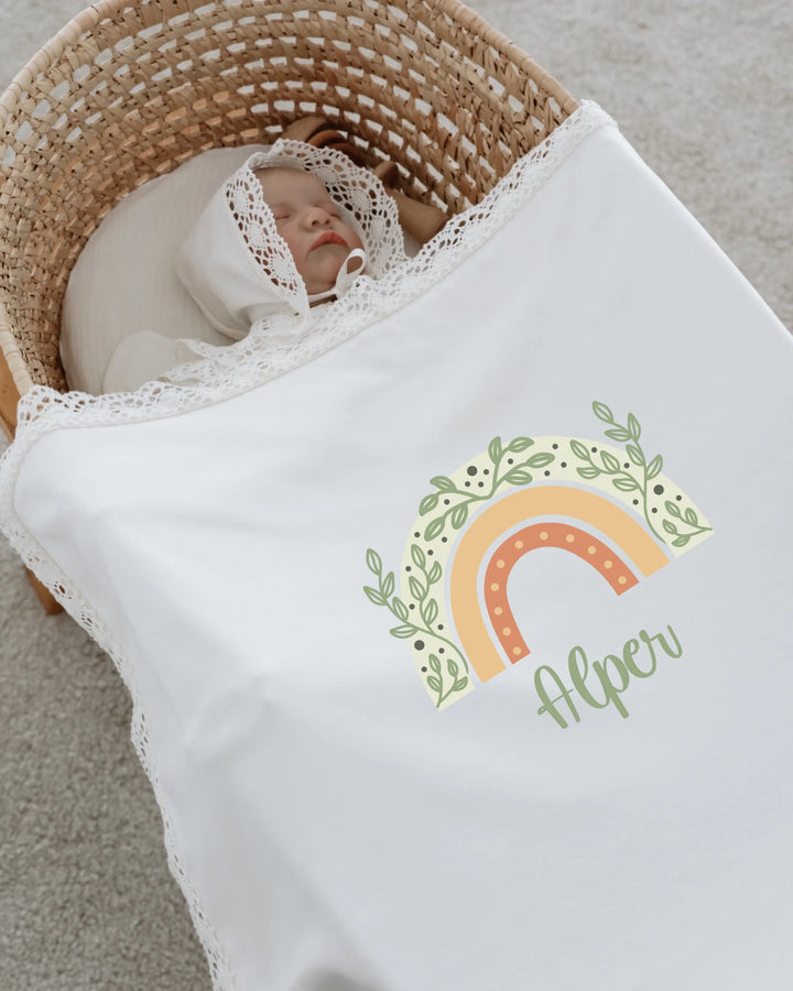 Baby wrapped in a white blanket with a colorful rainbow design and the name Alper, resting in a cozy basket.