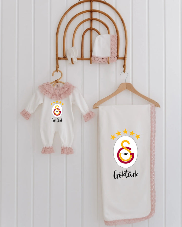 Galatasaray newborn set with logo, pink lace details, including romper and blanket, displayed on a wooden hanger.
