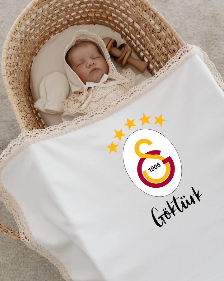 Newborn sleeping in a basket, wrapped in a personalized blanket with Galatasaray logo and name 'Göktürk'.