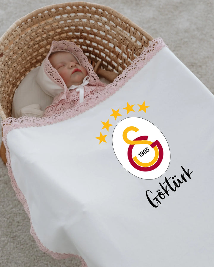 Newborn girl swaddled in a basket with Galatasaray logo and personalized name, showcasing a stylish baby outfit.