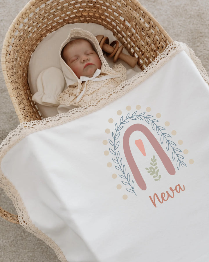 Newborn baby girl sleeping in a basket, wrapped in a personalized blanket with pastel rainbow design and name 'Neva'.