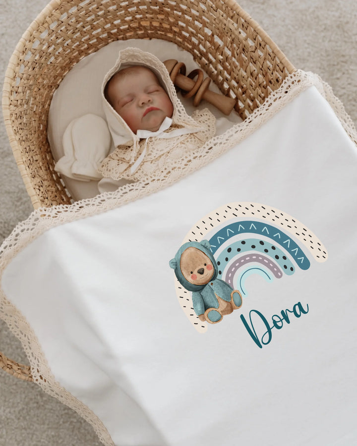 Newborn baby sleeping in a basket, wrapped in a personalized blanket featuring a rainbow and teddy bear design.