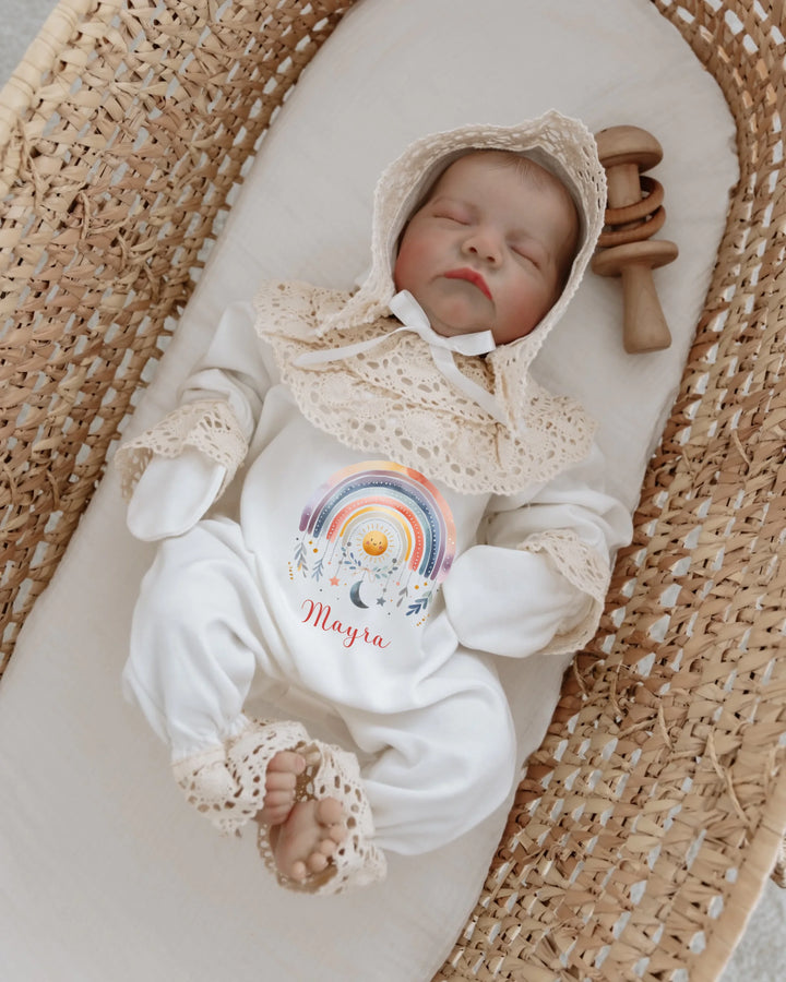 Newborn girl in a colorful rainbow romper with lace details, personalized with the name Mayra, resting in a woven basket.