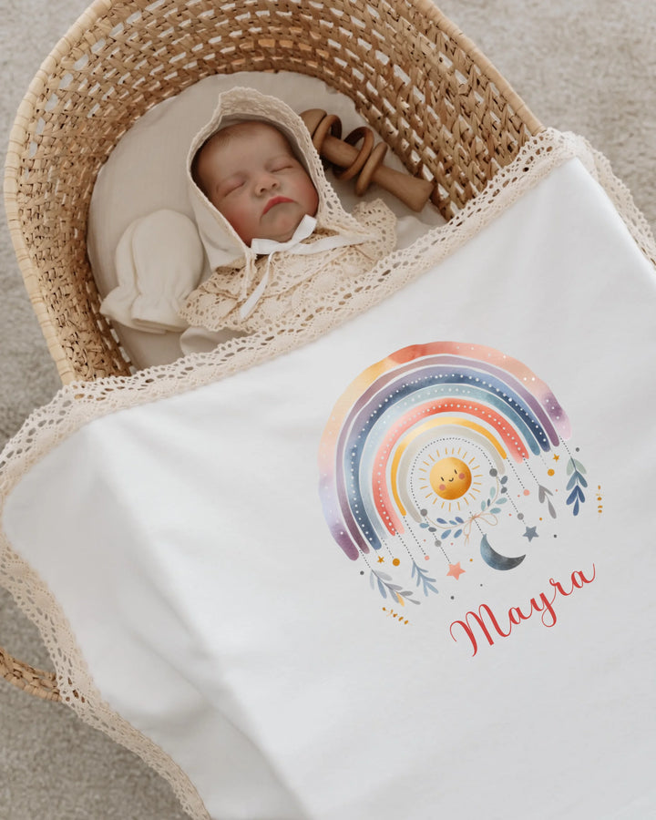 Newborn girl in cozy basket wrapped in personalized rainbow blanket with name Mayra.
