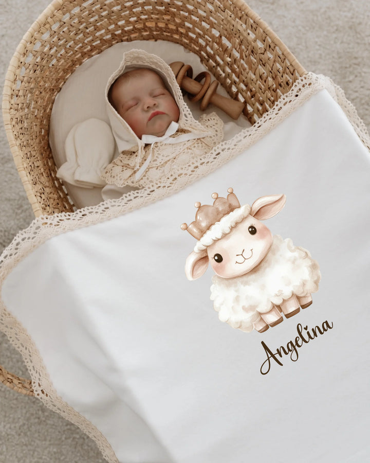 Newborn baby girl sleeping in a basket, covered with blanket featuring a crown sheep design and personalized name.