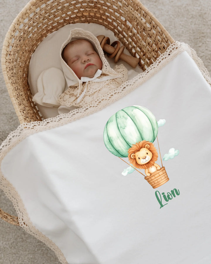 Baby girl in a basket with a lion hot air balloon design on her blanket, wearing a lace bonnet.