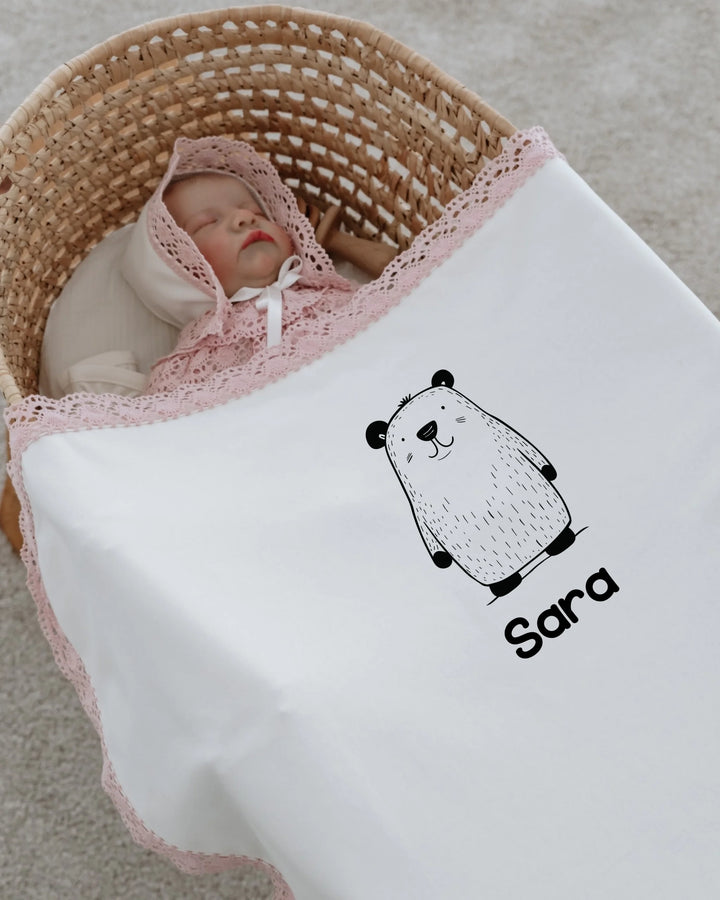 Newborn baby wrapped in a personalized blanket with a bear illustration and lace trim.