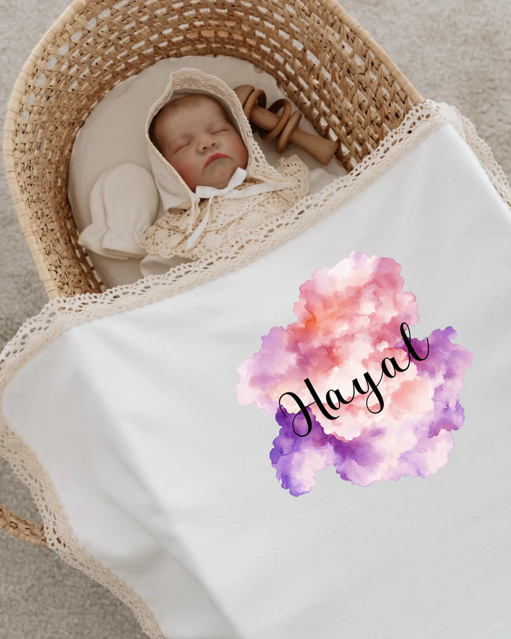 Baby sleeping peacefully in a basket, wrapped in a personalized pastel cloud blanket with the name 'Hayal'.