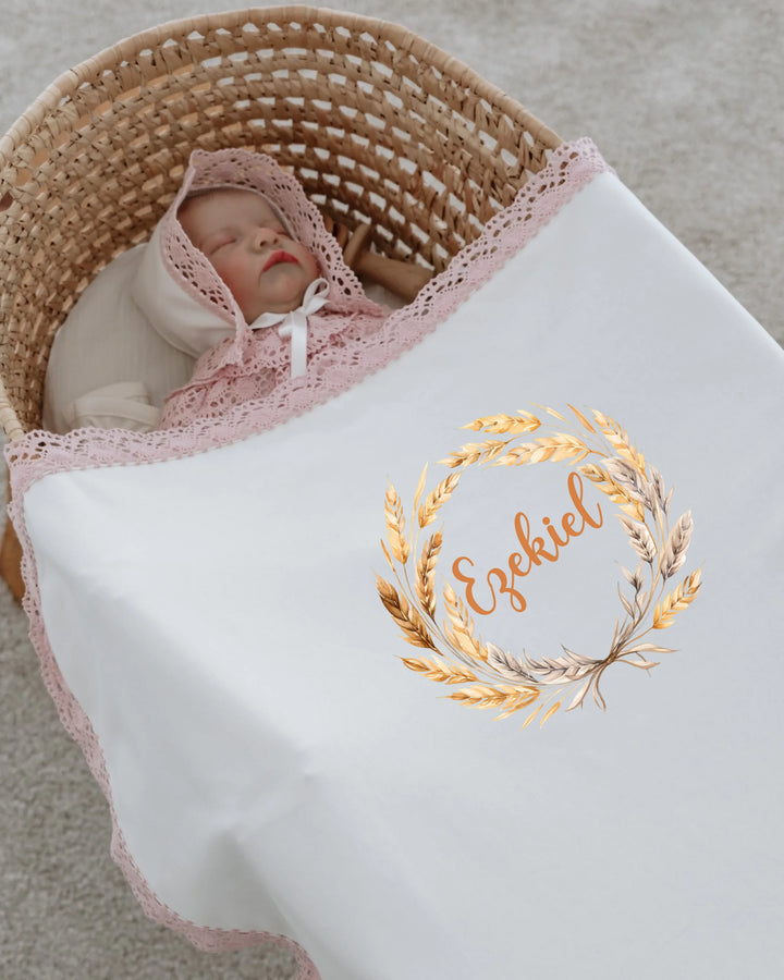 Baby wrapped in a stylish white blanket with a floral design and the name 'Ezekiel' in gold, nestled in a basket.