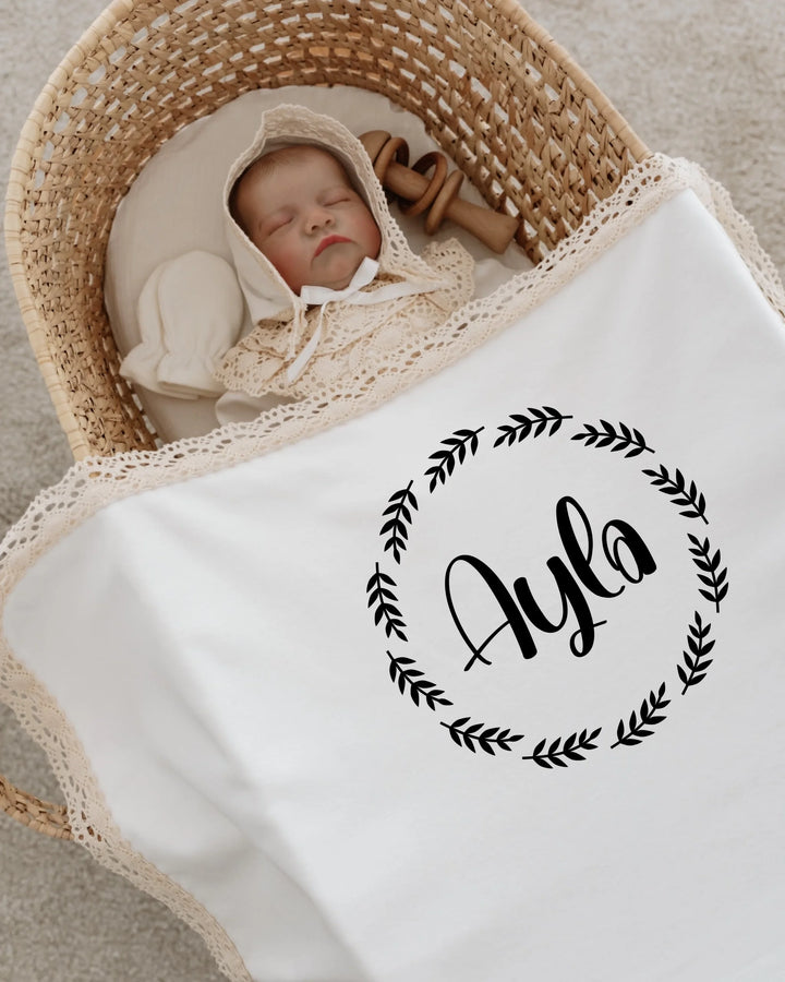 Newborn sleeping in a wicker basket wrapped in a personalized blanket featuring the name Ayla surrounded by a floral design.