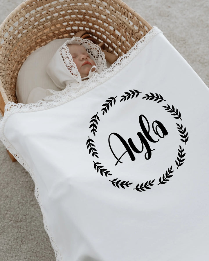 Newborn girl wrapped in white blanket with lace, featuring the name Ayla in a circular floral design.