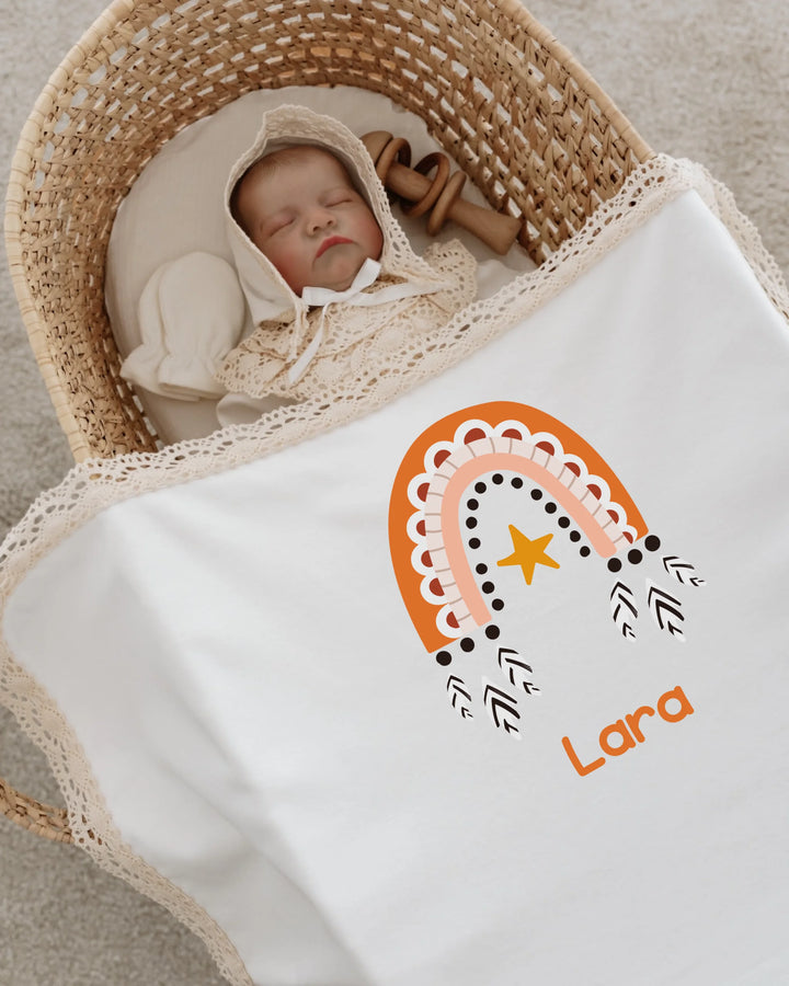 Pasgeboren meisje in een mand met een gepersonaliseerd dekentje met boho regenboogdesign en naam Lara.