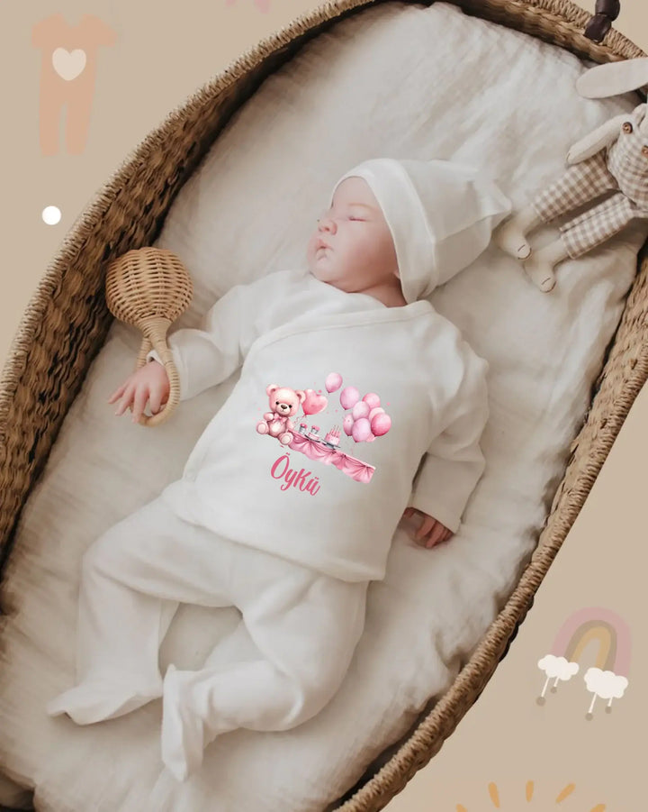 Baby in white personalized outfit featuring teddy bear and balloons, resting comfortably in a cozy basket.