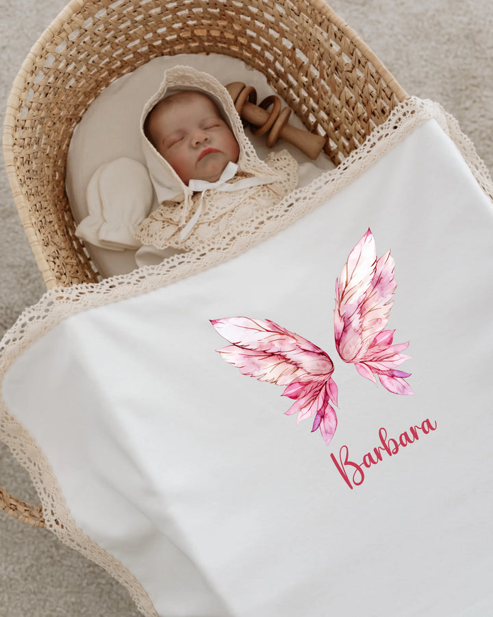 Newborn girl sleeping in a basket with personalized blanket featuring pink angel wings and name Barbara.